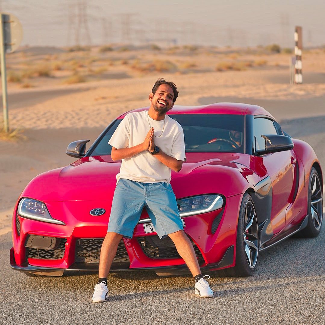 Mr. Indian Hacker poses confidently next to a striking red sports car, embodying a blend of charisma and automotive enthusiasm.