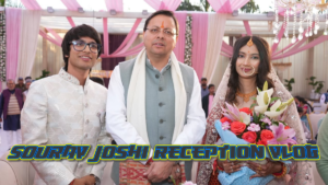 Three people (Sourav Joshi, sourav joshi wife and, Pushkar Singh Dhami, ) pose at an event under pink and white drapes. A man in glasses smiles, another wears a traditional suit, and a sourav joshi's wife (avantika bhatt) holds a bouquet, looking delighted.